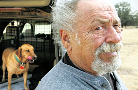 Cropped image of Jim Harrison outdoors, standing in front of a van with a brown dog.